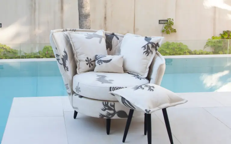Armchair upholstered in Quetzal fabric, a light-coloured textile with decorative patterns in grey tones, accompanied by matching cushions beside an outdoor pool.