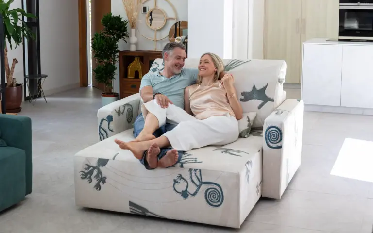 Couple sitting on a chaise longue sofa upholstered in Quetzal fabric, a light-coloured textile with decorative patterns in blue and grey tones, in a modern and bright living room.