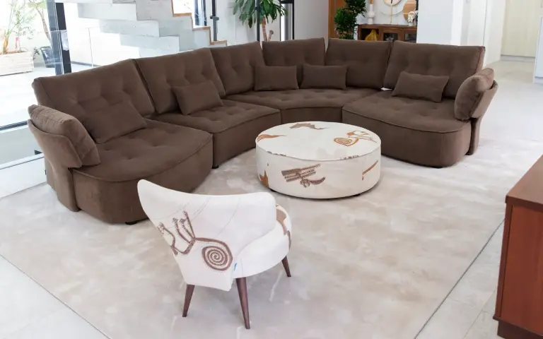 Chair and footstool upholstered in Quetzal fabric, a light-coloured textile with decorative patterns in earthy tones, combined with a brown modular sofa in a modern and bright living room.