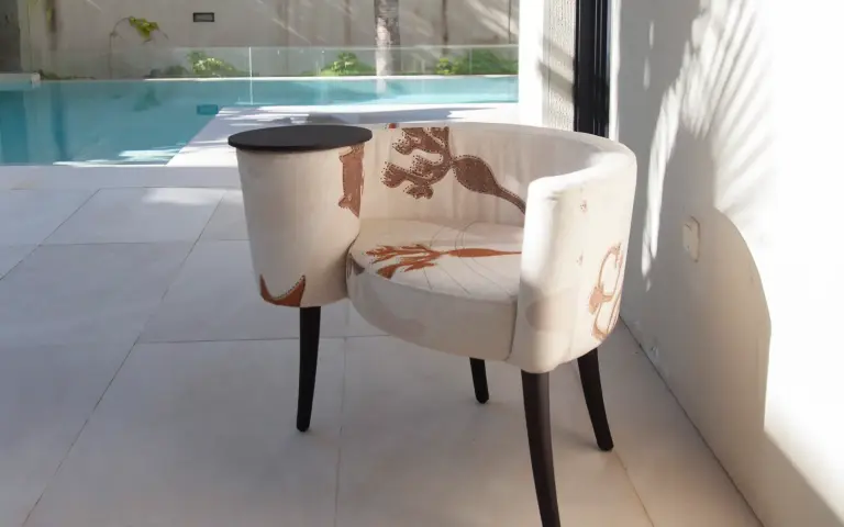 La Caracola armchair upholstered in Quetzal fabric, a light-coloured textile with decorative patterns in earthy tones, beside a pool in a bright and contemporary setting.