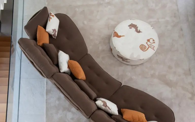 Top view of a brown modular sofa combined with a footstool upholstered in Quetzal fabric, a light-coloured textile with decorative patterns in earthy tones, in a contemporary living room.