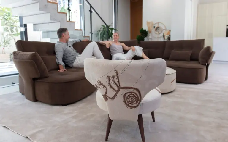 Chair upholstered in Quetzal fabric, a light-coloured textile with decorative patterns in earthy tones, combined with a brown modular sofa in a modern and bright living room.