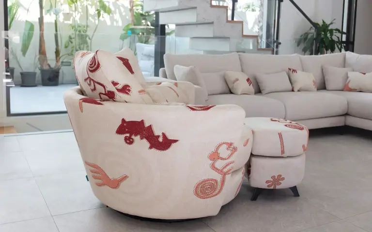 Armchair and footstool upholstered in Quetzal fabric, a light-coloured textile with decorative patterns in reddish and pink tones, combined with a light modular sofa in a modern and bright living room.